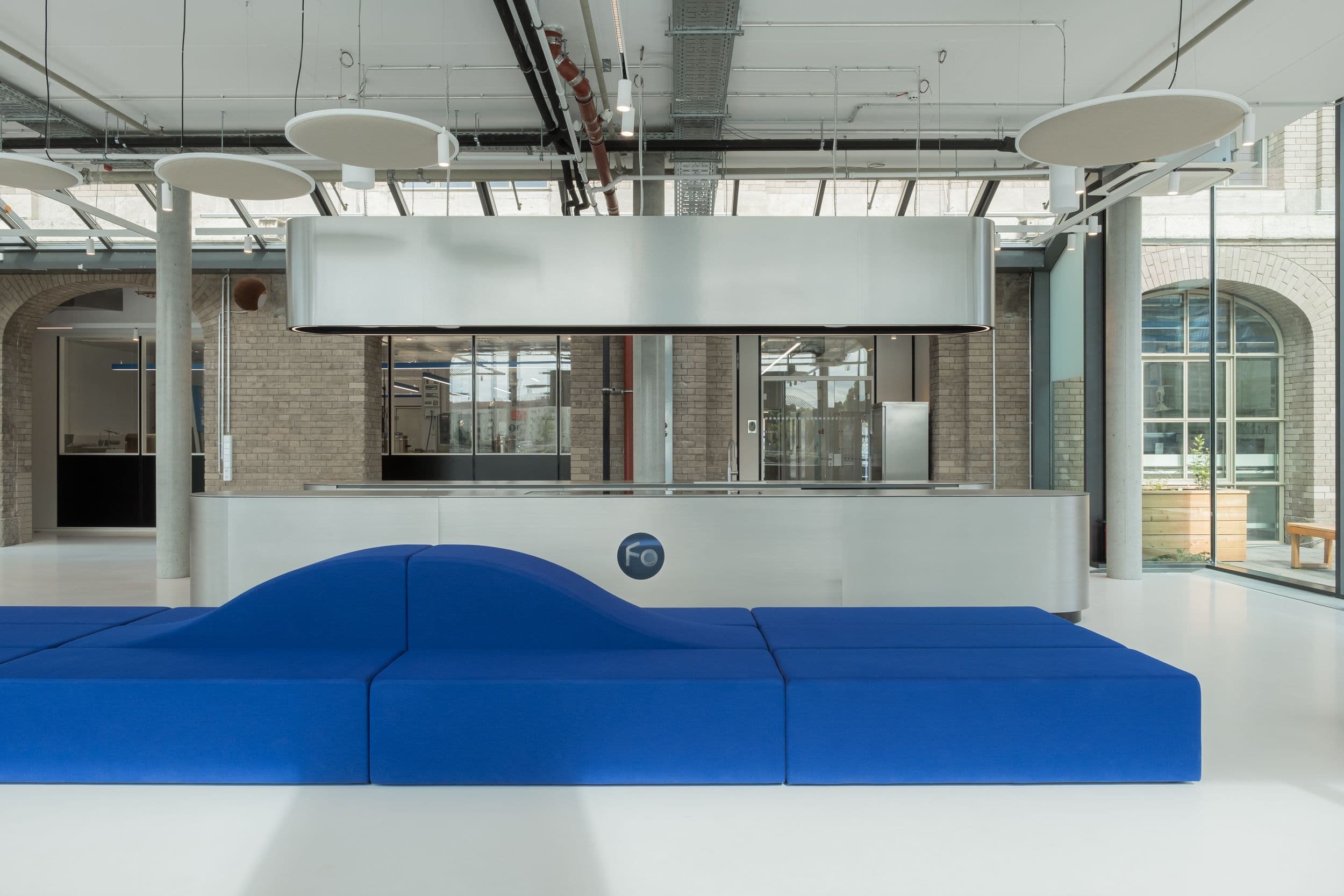 Offener Marktplatzbereich der Formo Zentrale Berlin mit blauer modularer Sitzlandschaft, geschwungener Edelstahlkücheninsel, Logofolie auf der Glasfassade und Blick auf die Spree, Innenarchitektur Susanne Philippson