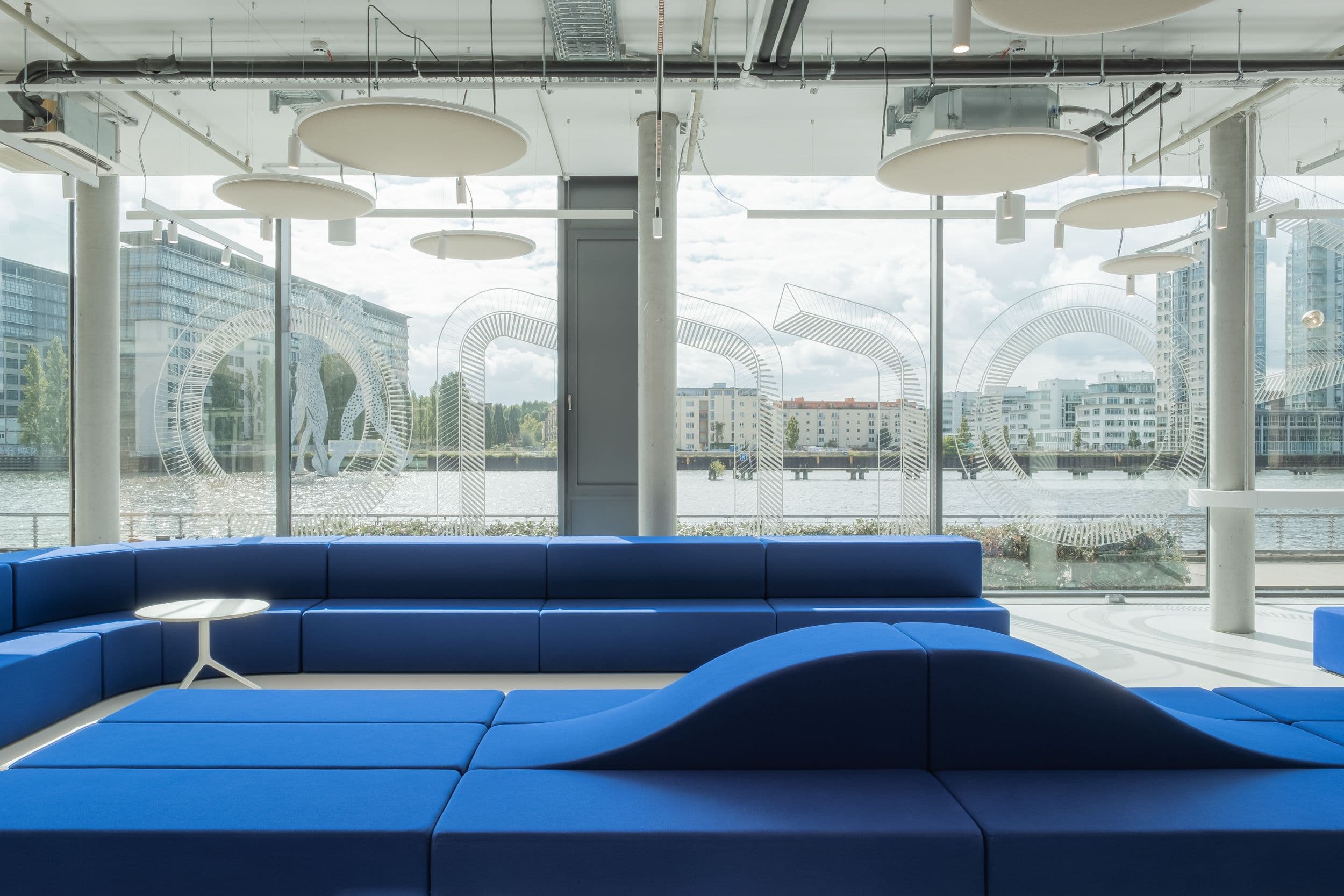 Offener Marktplatzbereich der Formo Zentrale Berlin mit blauer modularer Sitzlandschaft, geschwungener Edelstahlkücheninsel, Logofolie auf der Glasfassade und Blick auf die Spree, Innenarchitektur Susanne Philippson