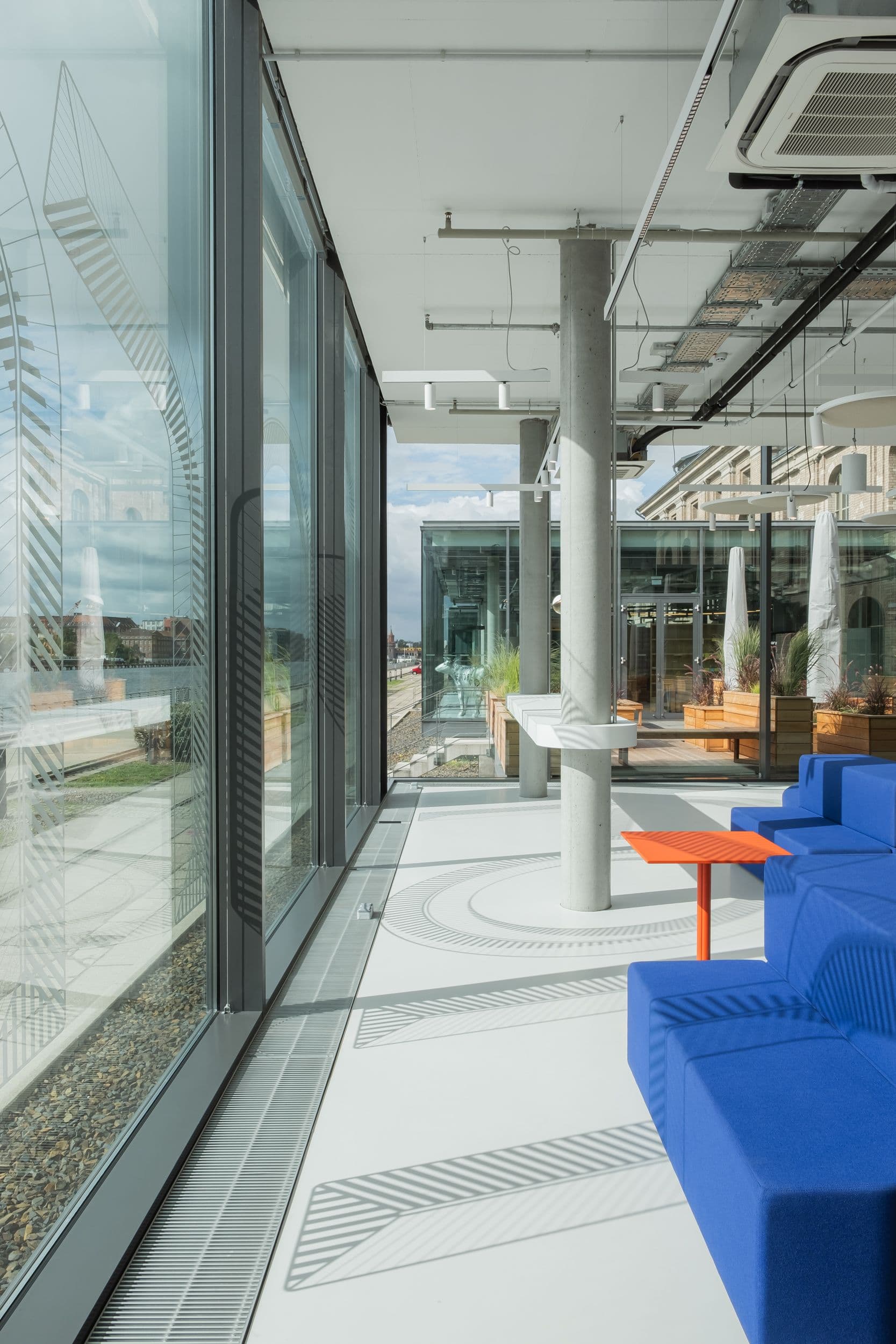 Offener Marktplatzbereich der Formo Zentrale Berlin mit blauer modularer Sitzlandschaft, geschwungener Edelstahlkücheninsel, Logofolie auf der Glasfassade und Blick auf die Spree, Innenarchitektur Susanne Philippson