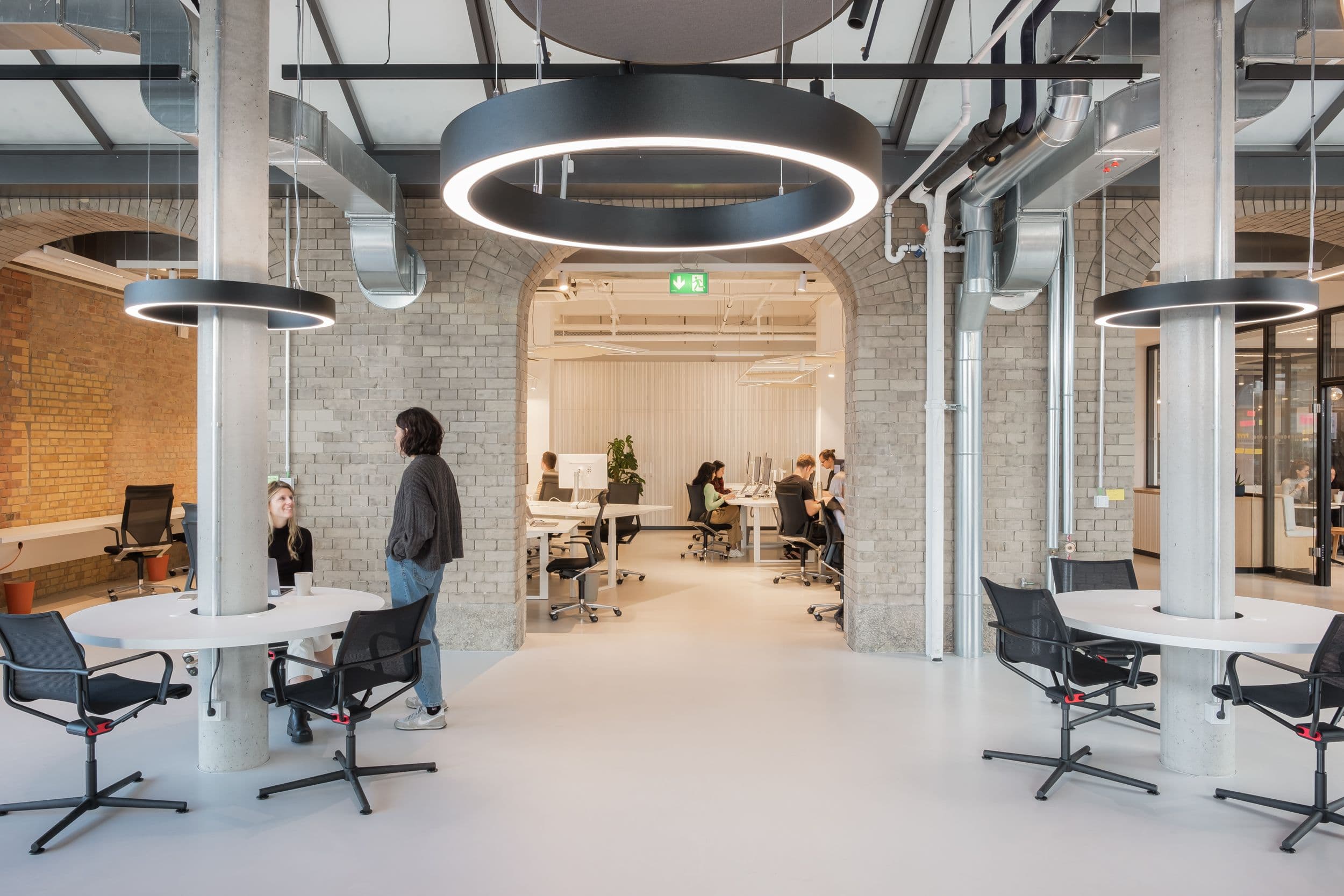 Offener Arbeitsbereich der Formo Zentrale Berlin mit Backsteinbögen, höhenverstellbaren Schreibtischen, quadratischen Akustik-Lichtdecken und Blick auf den zentralen Kollaborationsbereich, Innenarchitektur Susanne Philippson