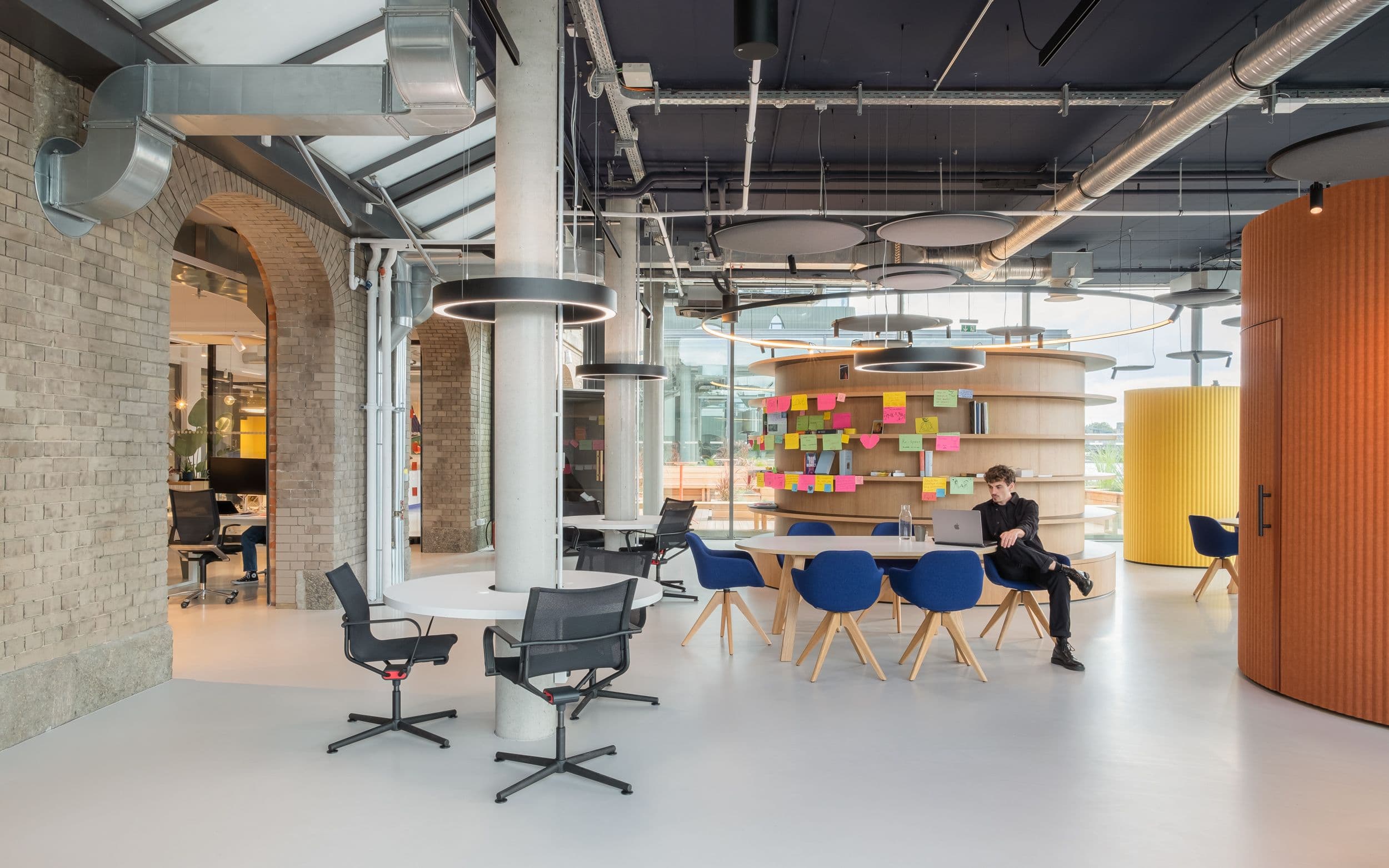 Offener Kollaborationsbereich der Formo Zentrale Berlin mit rundem Holzregal, farbigen Akustikzylindern in Gelb und Terrakotta, blauen Schalenstühlen und Blick auf die Spree, Innenarchitektur Susanne Philippson