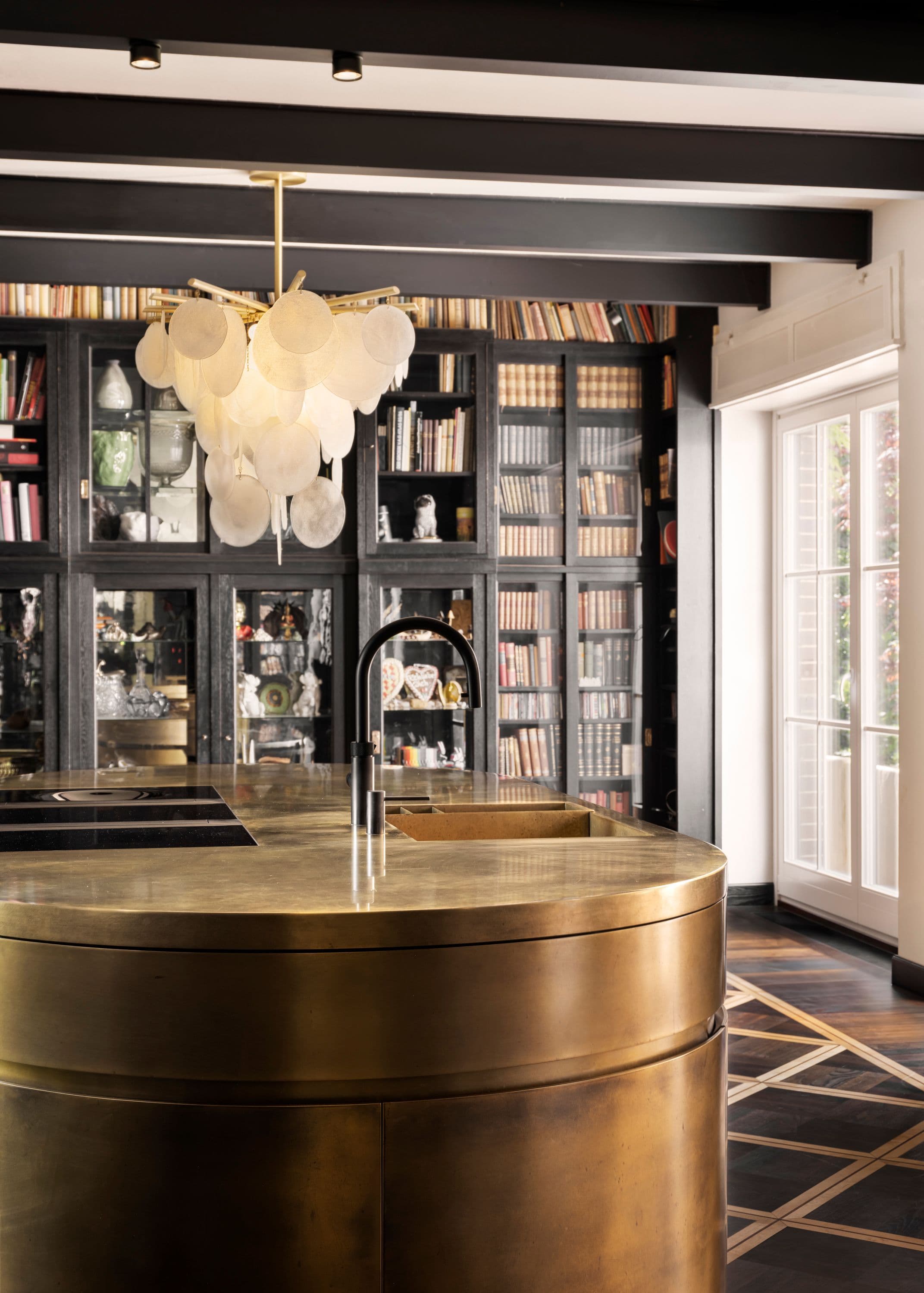 Bibliotheksküche eines Privathauses mit ovaler Bronzeküche, maßgefertigtem Tafelparkett mit Messingintarsien, dunklen Einbauten mit Spiegelglas und Perlmuttpendelleuchte vor raumhoher Bücherwand, Innenarchitektur Susanne Philippson