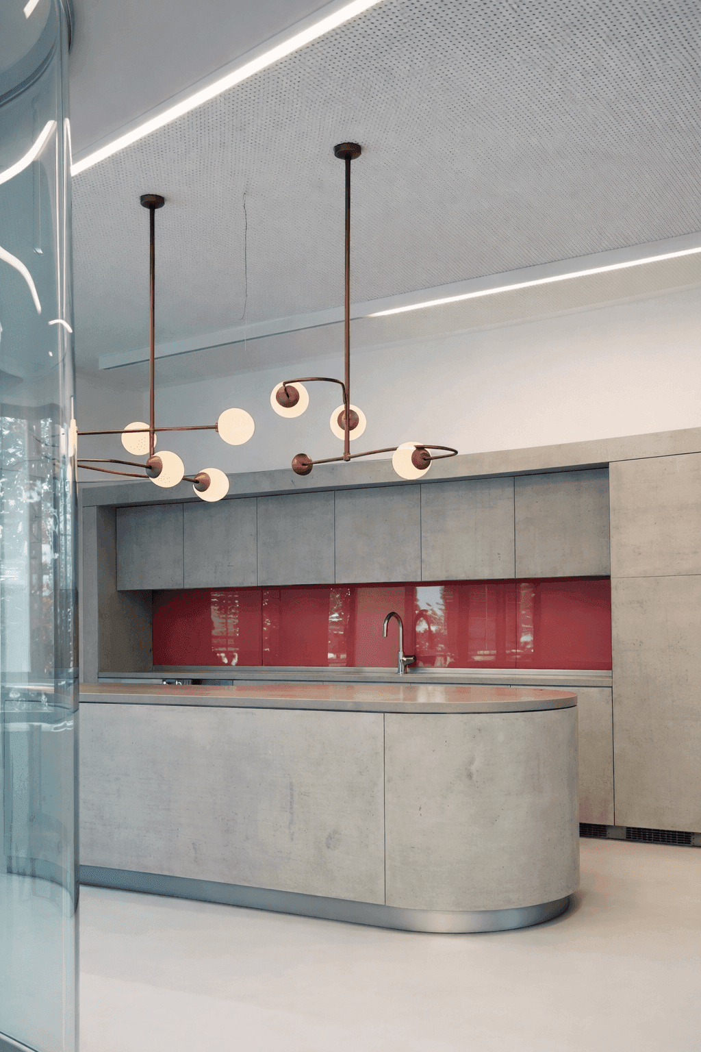Küchenbereich mit gerundetem Küchenblock in Betonoptik, rotem Glasrückwand-Akzent und Pendelleuchten mit Messinggestell im Büro Formo Frankfurt, Innenarchitektur Susanne Philippson
