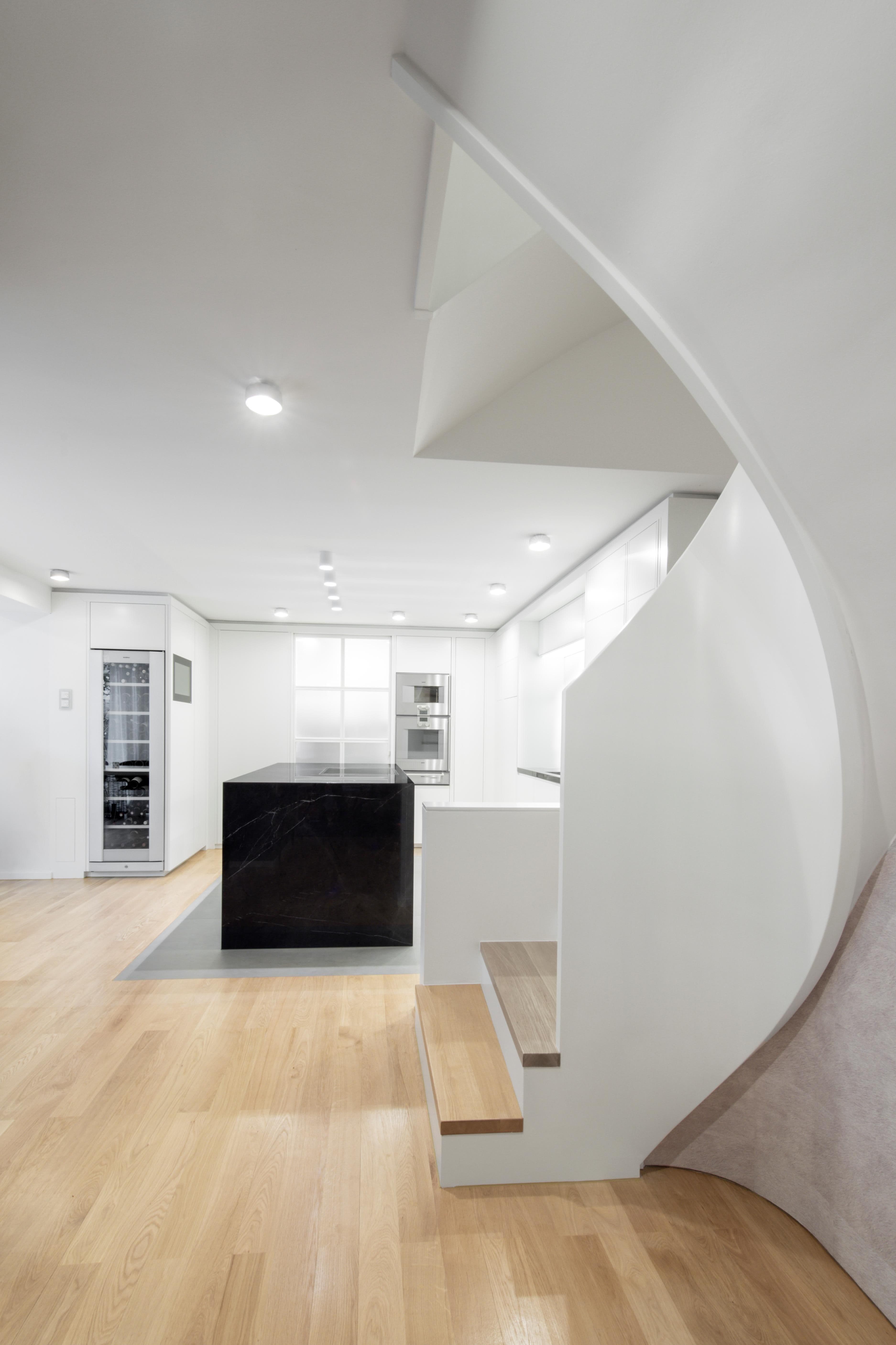 Offener Wohn- und Küchenbereich mit schwarzem Marmorinselblock, integrierten Gaggenau-Geräten, weißen grifflosen Einbauten und geschwungener Treppe mit Holzstufen in einer Privatwohnung, Innenarchitektur Susanne Philippson