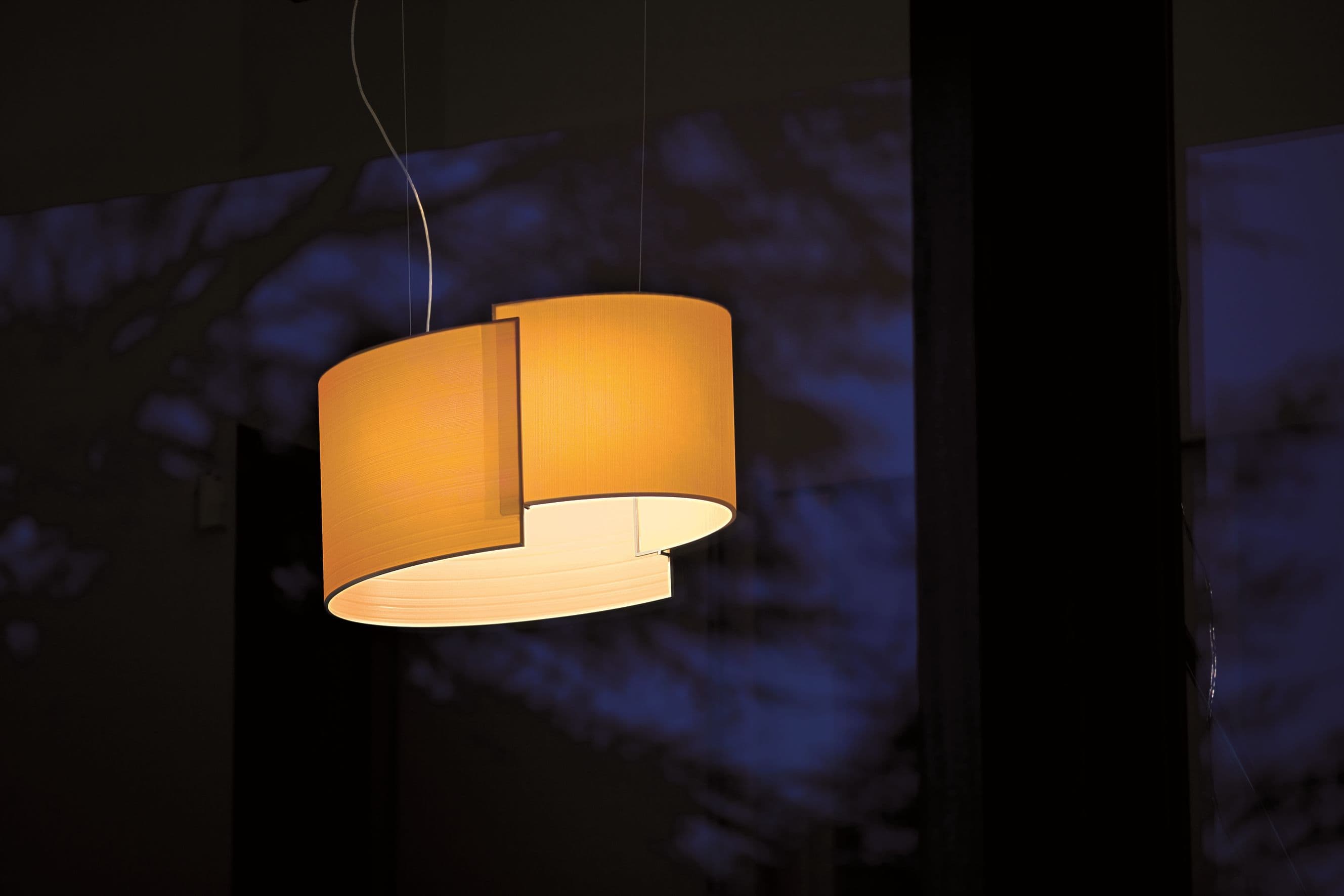 Joiin Pendelleuchte von Pallucco in Orange mit zwei versetzt kombinierten gebogenen Schirmflächen und warmem Lichtschein vor blauem Nachtambiente, Leuchtendesign Susanne Philippson