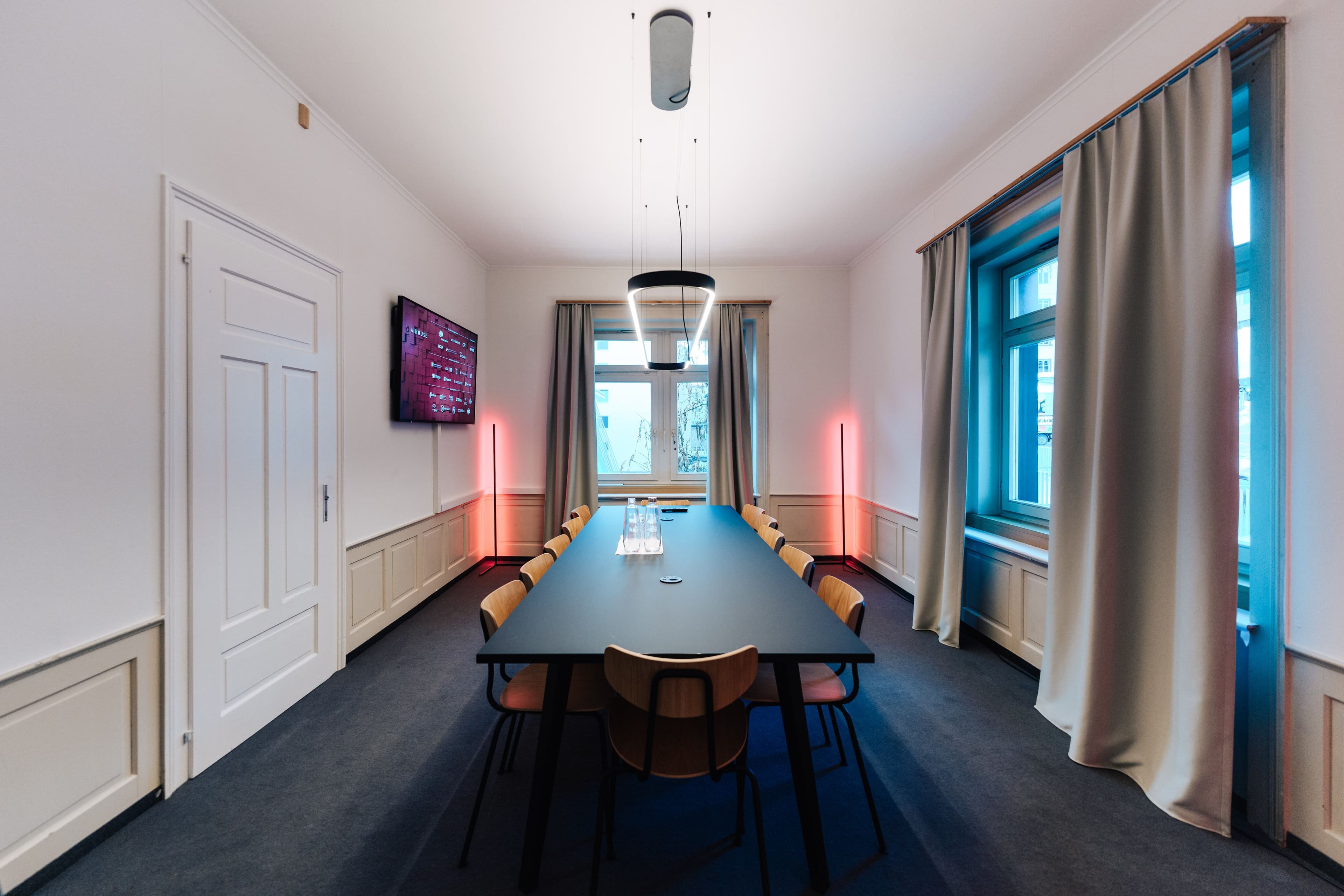Besprechungsraum im Altbau mit dunklem Konferenztisch, Holzstühlen, ringförmiger Pendelleuchte und schlanken Stehleuchten mit rotem Lichtakzent vor historischer Wandvertäfelung, Innenarchitektur Susanne Philippson