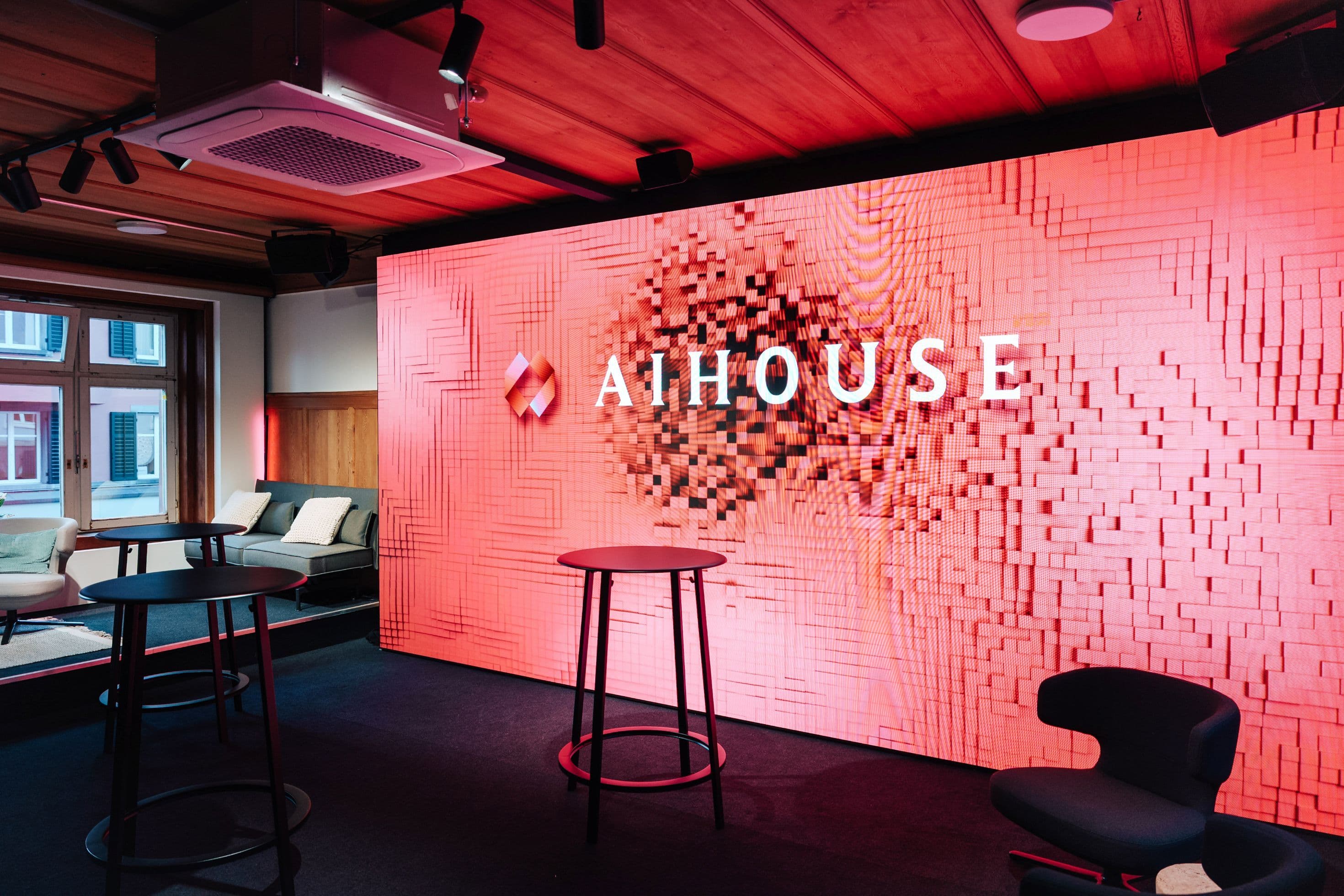 Eventbereich mit großformatigem LED-Display mit AI House-Branding, Stehtischen, Loungesessel und Sofas vor Altbaufenstern mit roter Akzentbeleuchtung, Innenarchitektur Susanne Philippson