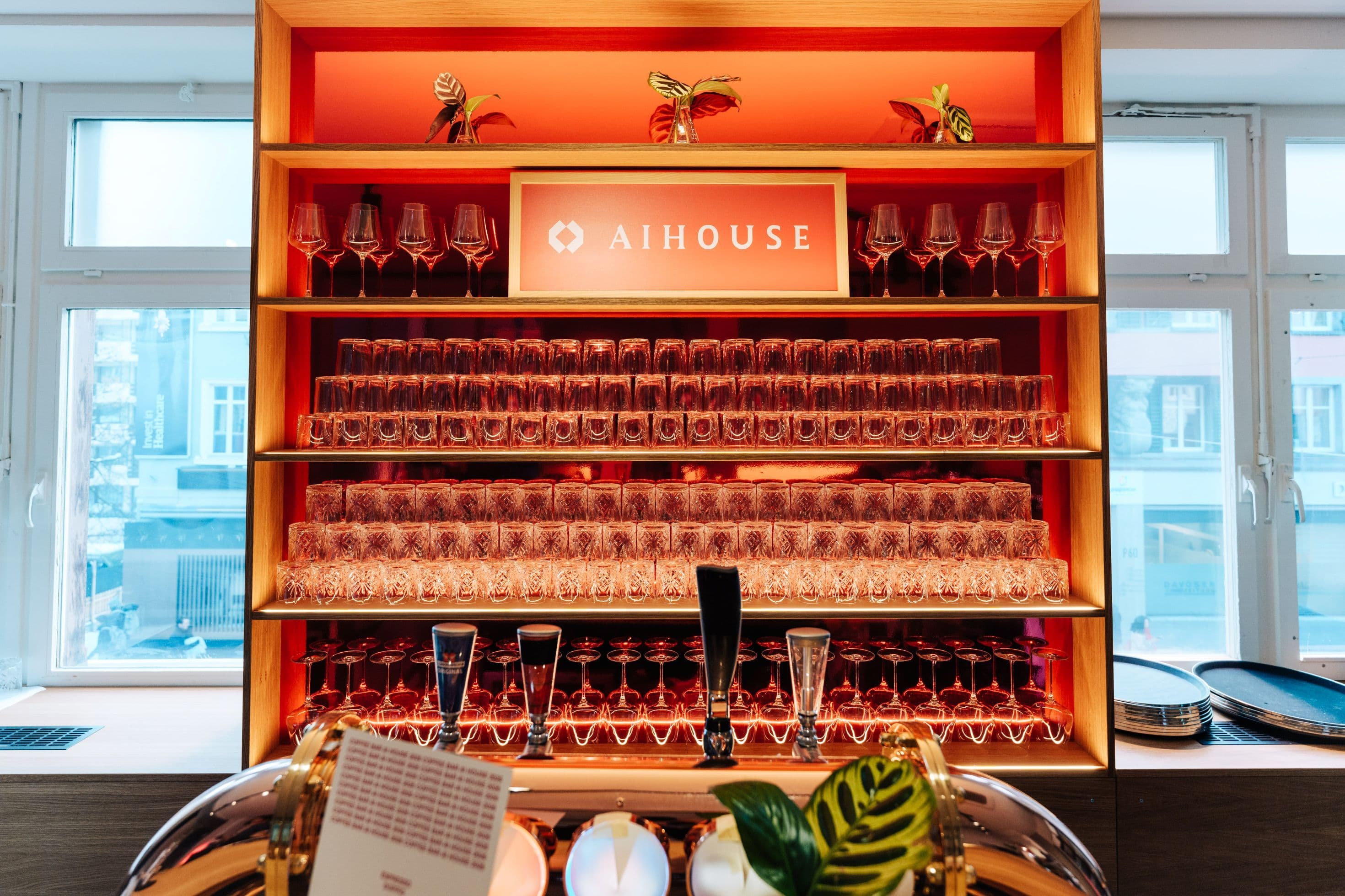 Barbereich mit umlaufender Holztheke, hinterbeleuchtetem Regalrückwand mit AI House-Branding und Glasbestückung sowie Natursteinverkleidung an der Thekenfront, Innenarchitektur Susanne Philippson