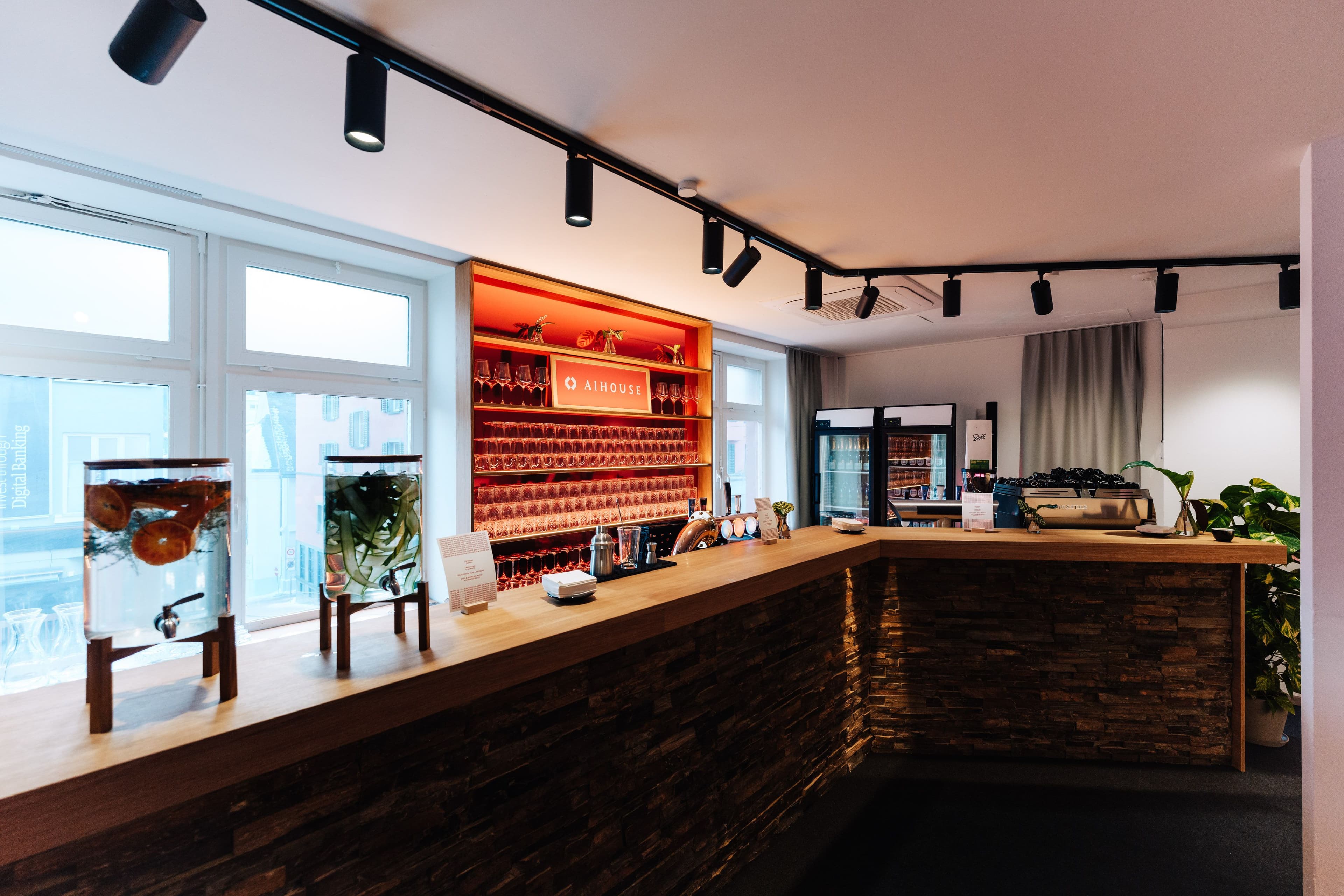 Barbereich mit umlaufender Holztheke, hinterbeleuchtetem Regalrückwand mit AI House-Branding und Glasbestückung sowie Natursteinverkleidung an der Thekenfront, Innenarchitektur Susanne Philippson