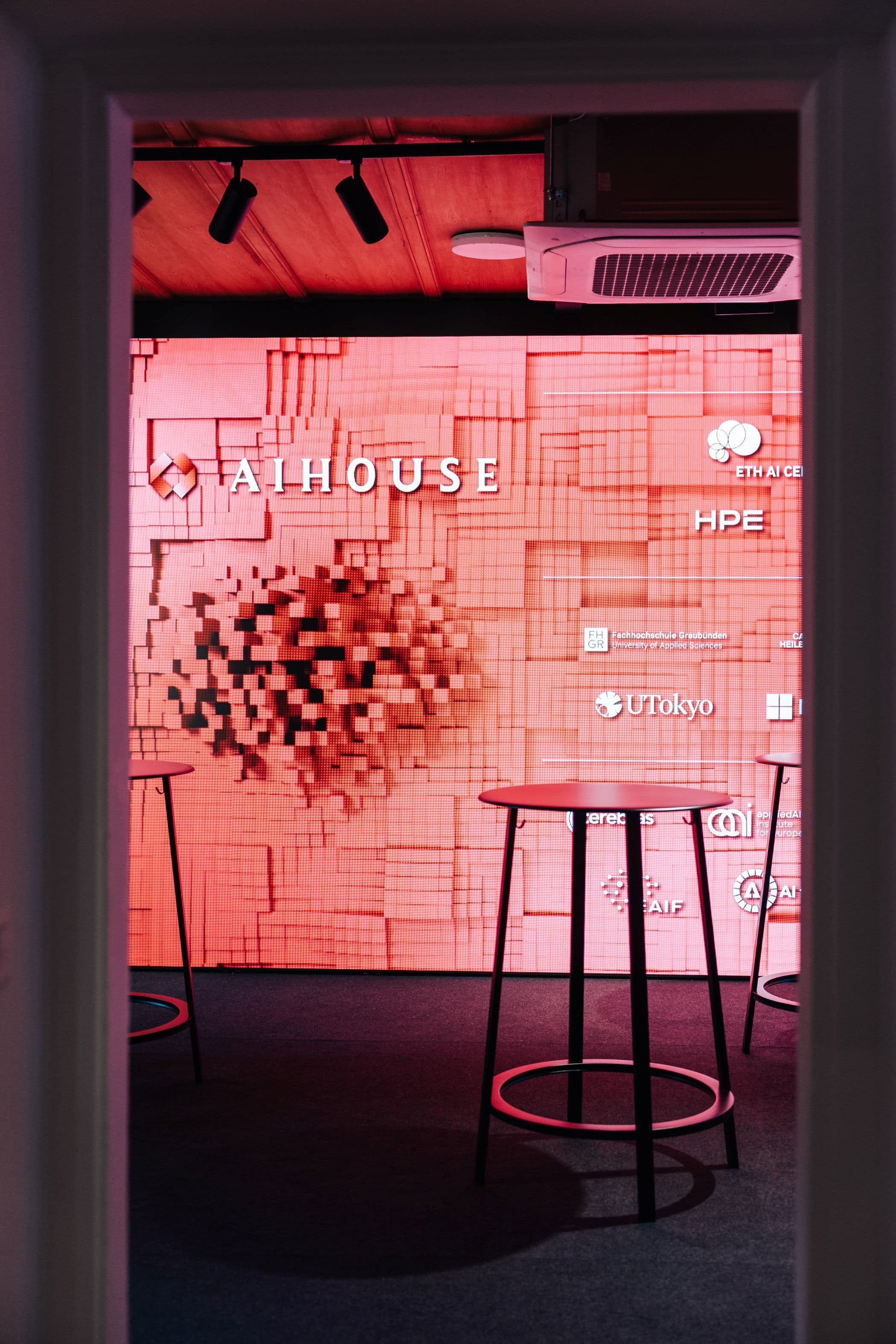 Eventbereich mit großformatigem LED-Display mit AI House-Branding, Stehtischen, Loungesessel und Sofas vor Altbaufenstern mit roter Akzentbeleuchtung, Innenarchitektur Susanne Philippson