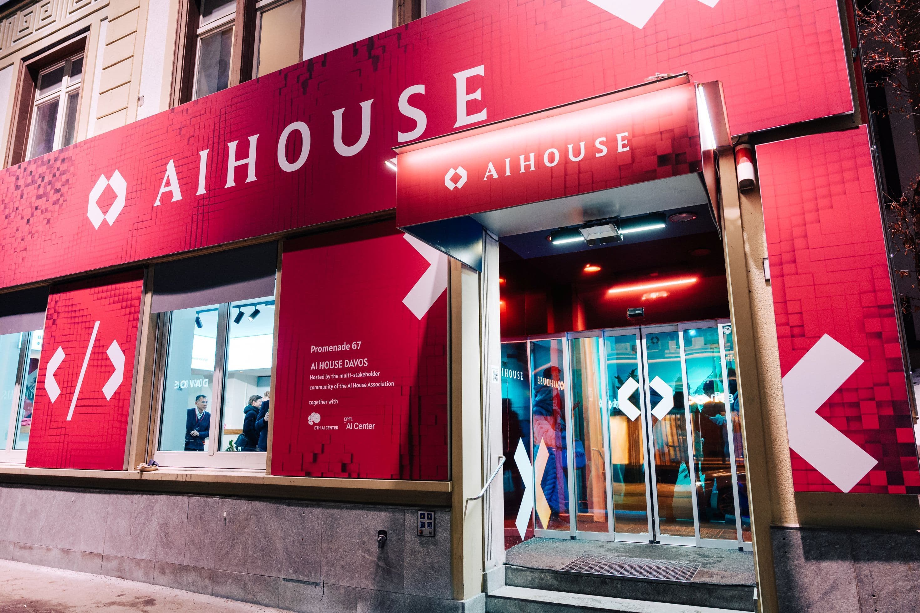 Nachtaufnahmen der AI House Davos Fassade an der Promenade mit großflächiger roter Markenbeschriftung, beleuchtetem Eingangsbereich und winterlicher Davoser Innenstadt, Innenarchitektur Susanne Philippson
