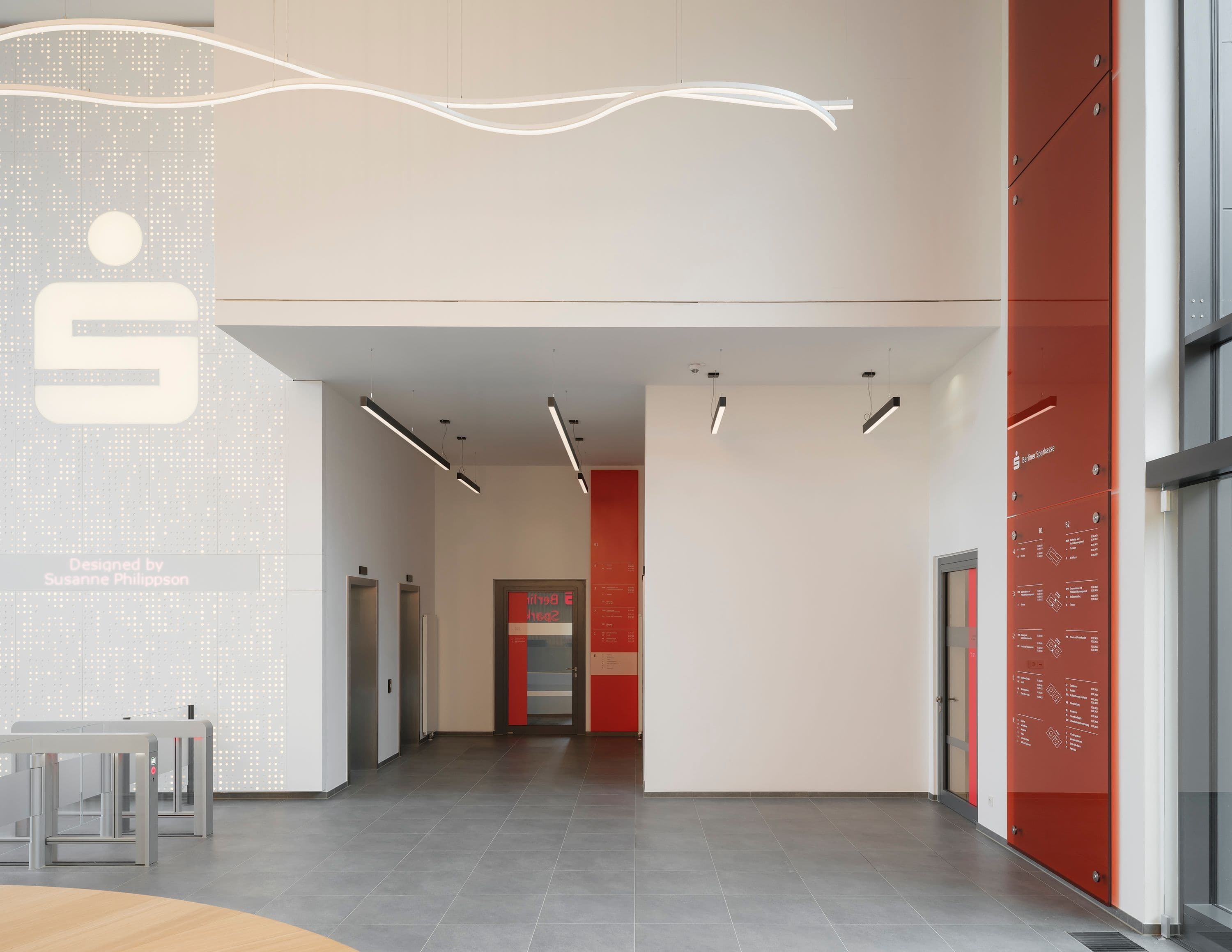 Eingangsbereich der Berliner Sparkasse Zentrale mit großformatigem Sparkassen-Logo auf perforierter Wandfolie, roter Orientierungsstele mit Geschossübersicht und geschwungener Lichtinstallation an der Decke, Signaletik Susanne Philippson