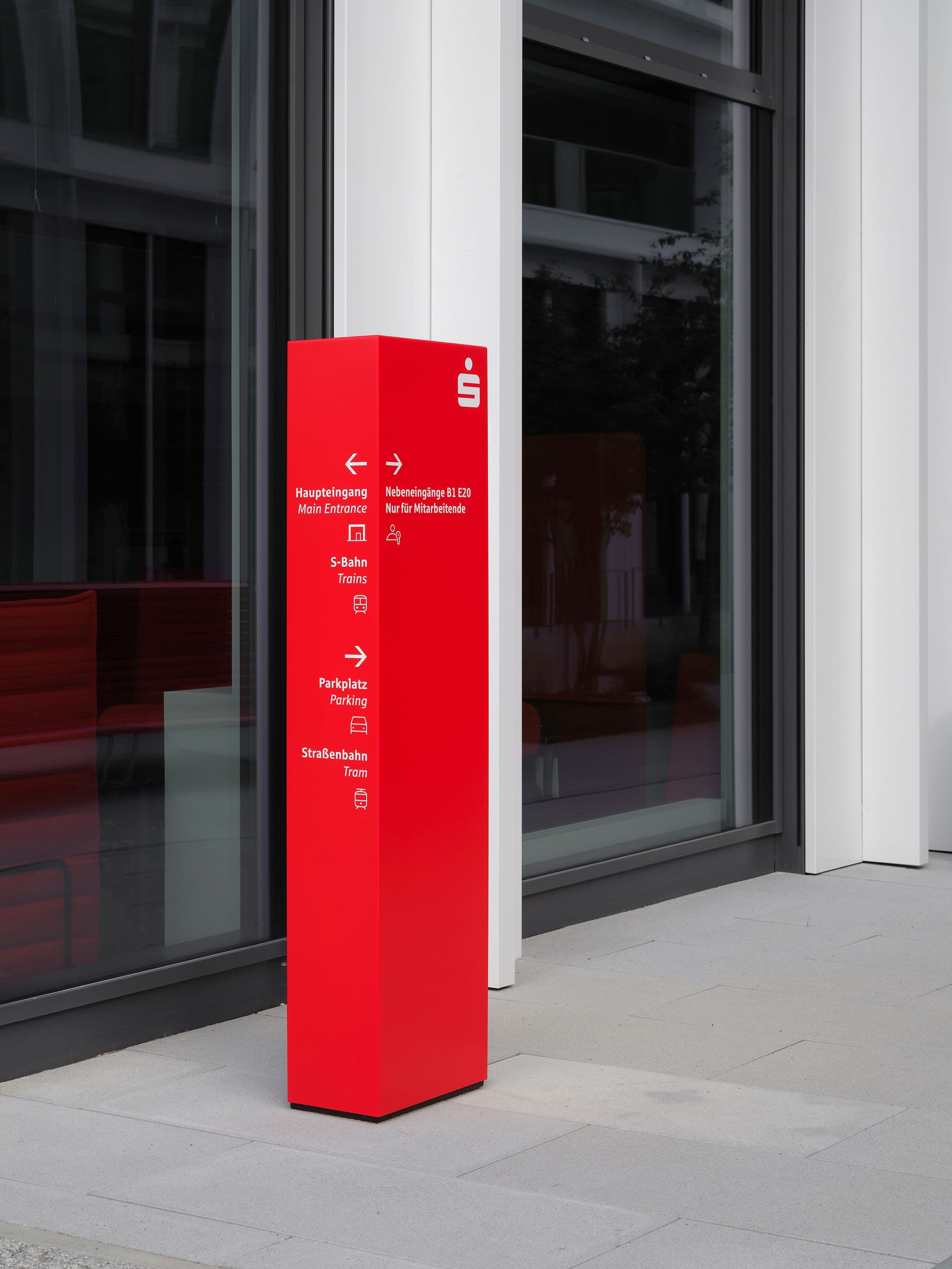 Außenbereich der Berliner Sparkasse Zentrale mit überdachtem Durchgang, begrüntem Innenhof und roter Wegweiserstele mit zweisprachiger Beschriftung zu Haupteingang, S-Bahn und Parkplatz, Signaletik Susanne Philippson