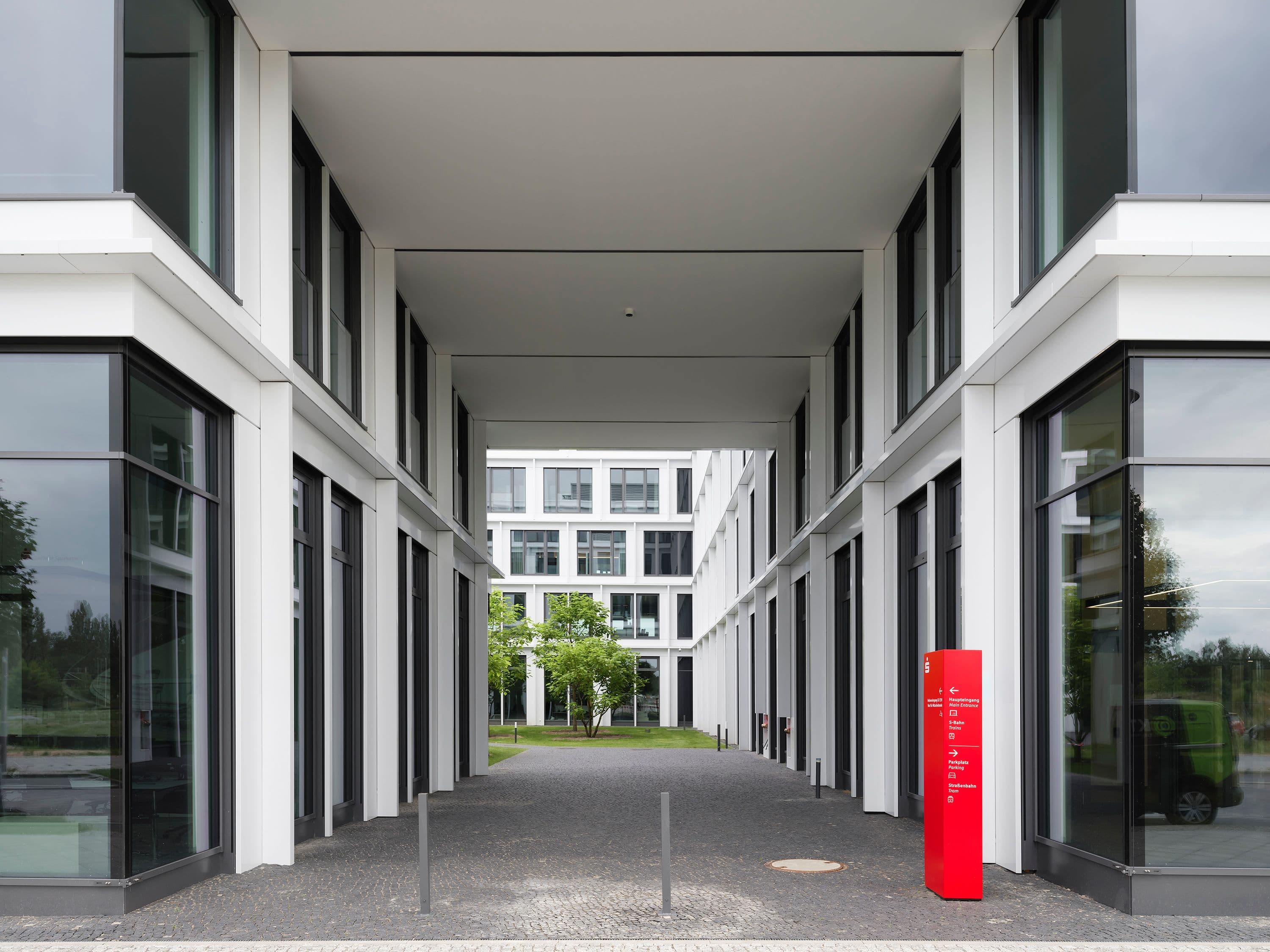 Außenbereich der Berliner Sparkasse Zentrale mit überdachtem Durchgang, begrüntem Innenhof und roter Wegweiserstele mit zweisprachiger Beschriftung zu Haupteingang, S-Bahn und Parkplatz, Signaletik Susanne Philippson