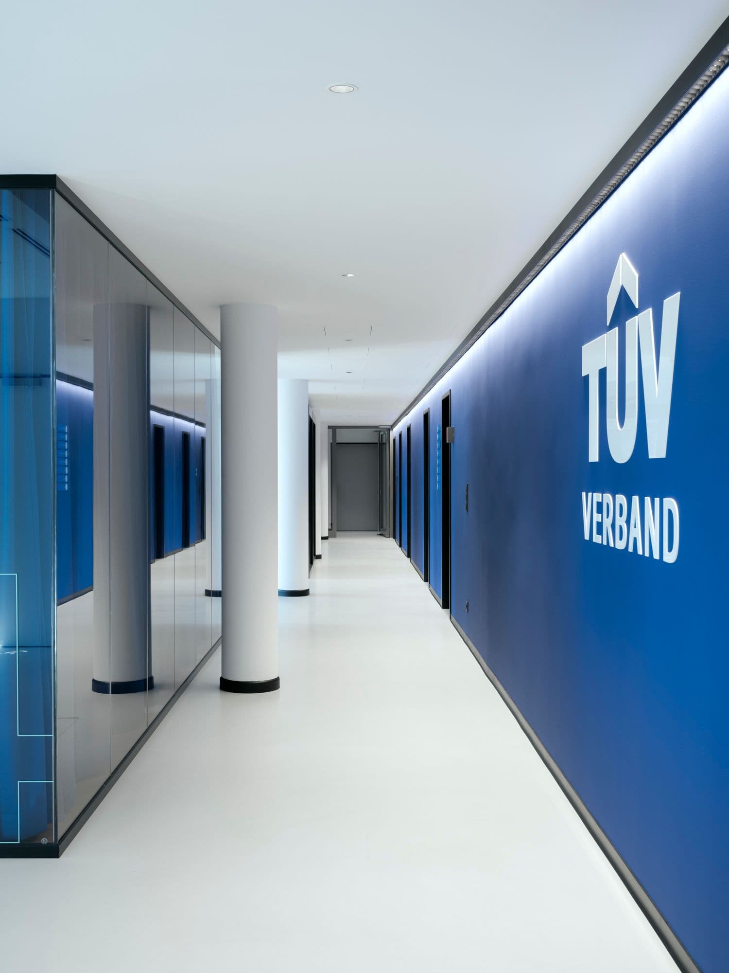 Langer Flur mit blauer TÜV-Verband-Markenwand, indirekter Wandbeleuchtung und Glastrennwänden im TÜV Verband Berlin