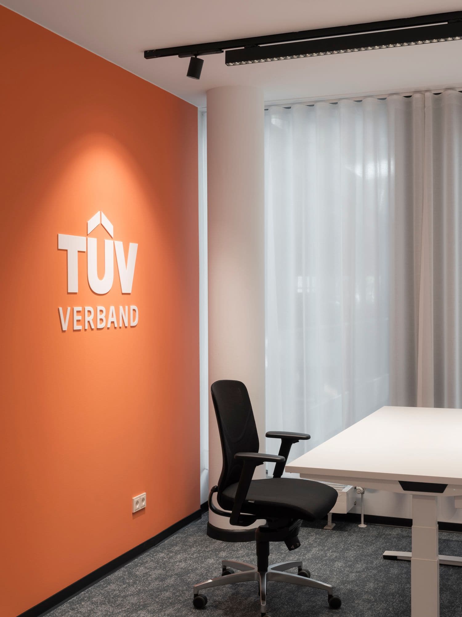 Einzelbüro mit orangefarbener Akzentwand, dreidimensionalem TÜV-Verband-Logo und Bürostuhl von Wilkhahn im TÜV Verband Berlin